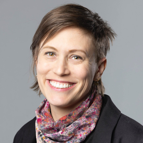 Smiling woman with short light brown hair wearing a black jacket and a colorful patterned scarf, posed against a gray background.
